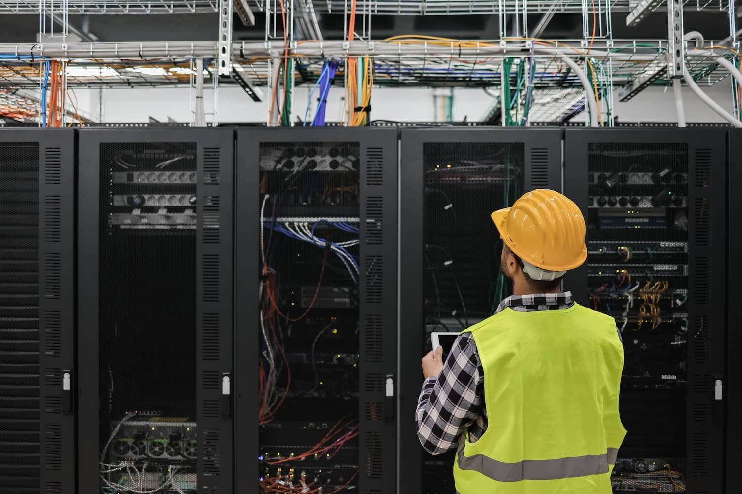 Man in safety vest and hard hat standing in front of a row of electrical equipment encased in black metal boxes.